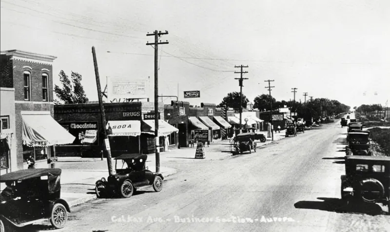 Historic photograph of the business section of Colfax Avenue in Aurora, Colorado, with storefront awnings, drugstore signs and early automobiles