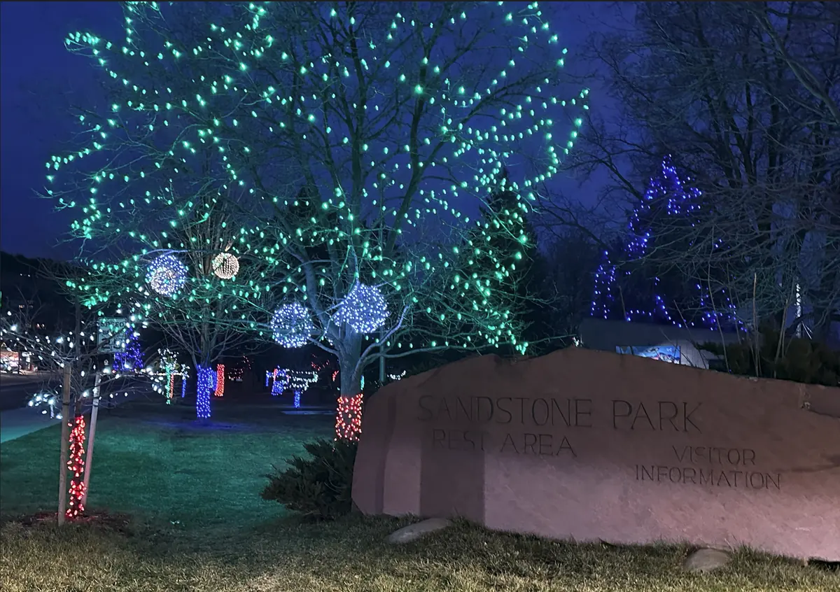 Sandstone Park in Lyons with a large tree filled with green lights, blue evergreens and lit spheres behind the park's sandstone sign