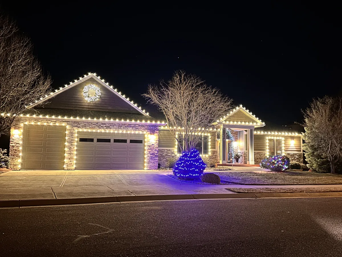 A stone-front ranch home lit for Christmas: warm white C9 bulbs along every roofline and around the two-car garage, a lit wreath on the front gable, an evergreen netted in blue, a multicolor shrub and a lit entry garland