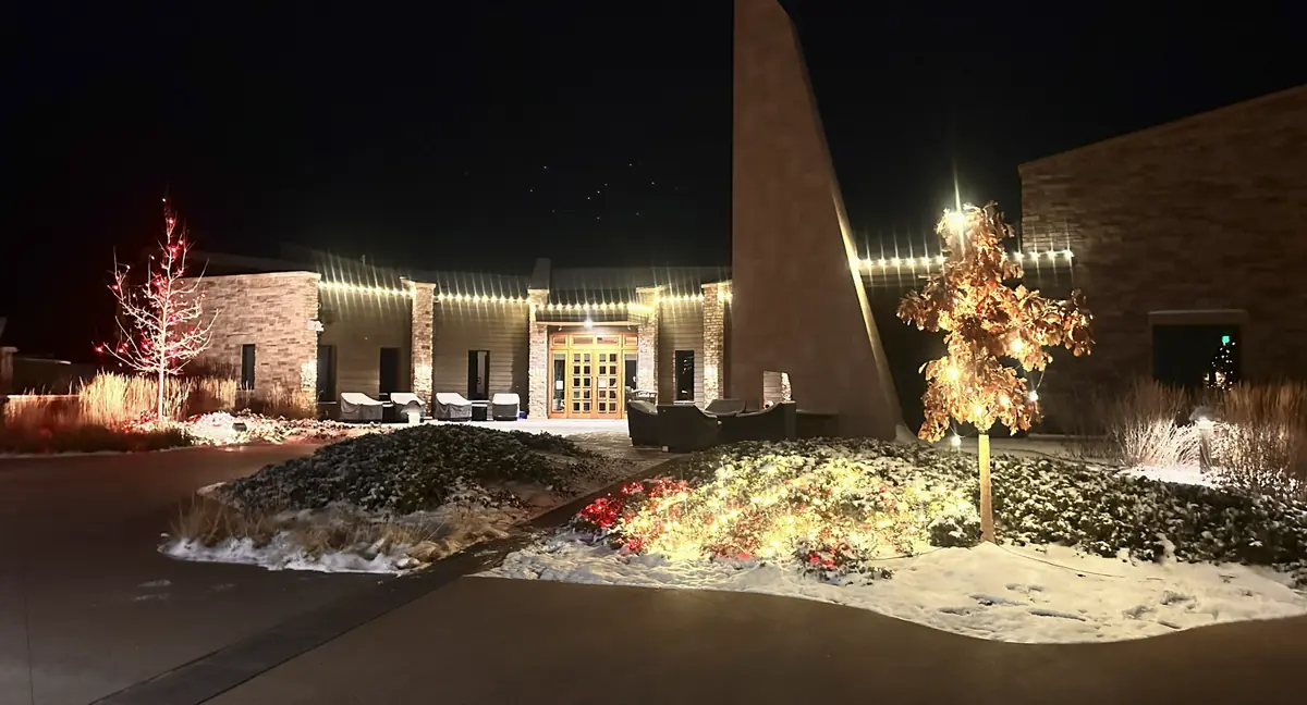 HOA clubhouse entrance with warm white roofline, an uplit ornamental tree and lit beds under snow