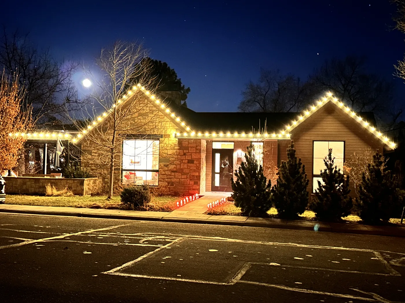 A Lafayette brick ranch outlined in warm white bulbs under a full moon, with lit evergreens along the front