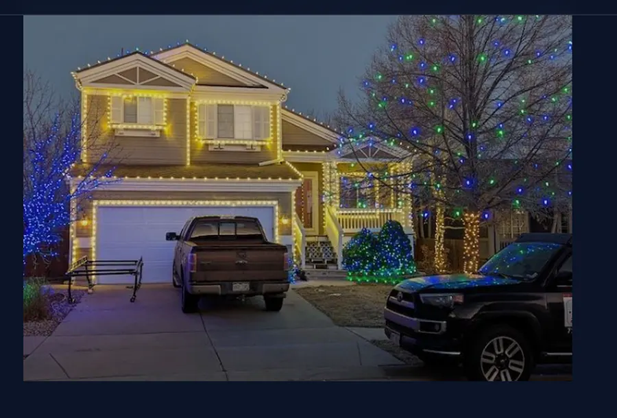 Two-story home outlined in warm white C9 bulbs along eaves, gables and dormers, with blue and green lit trees across the front yard