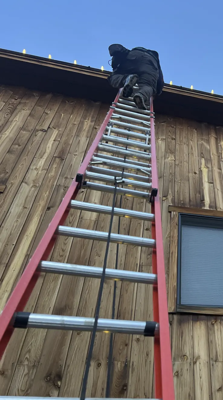 An FHL installer on an extension ladder clipping warm white bulbs along a roofline